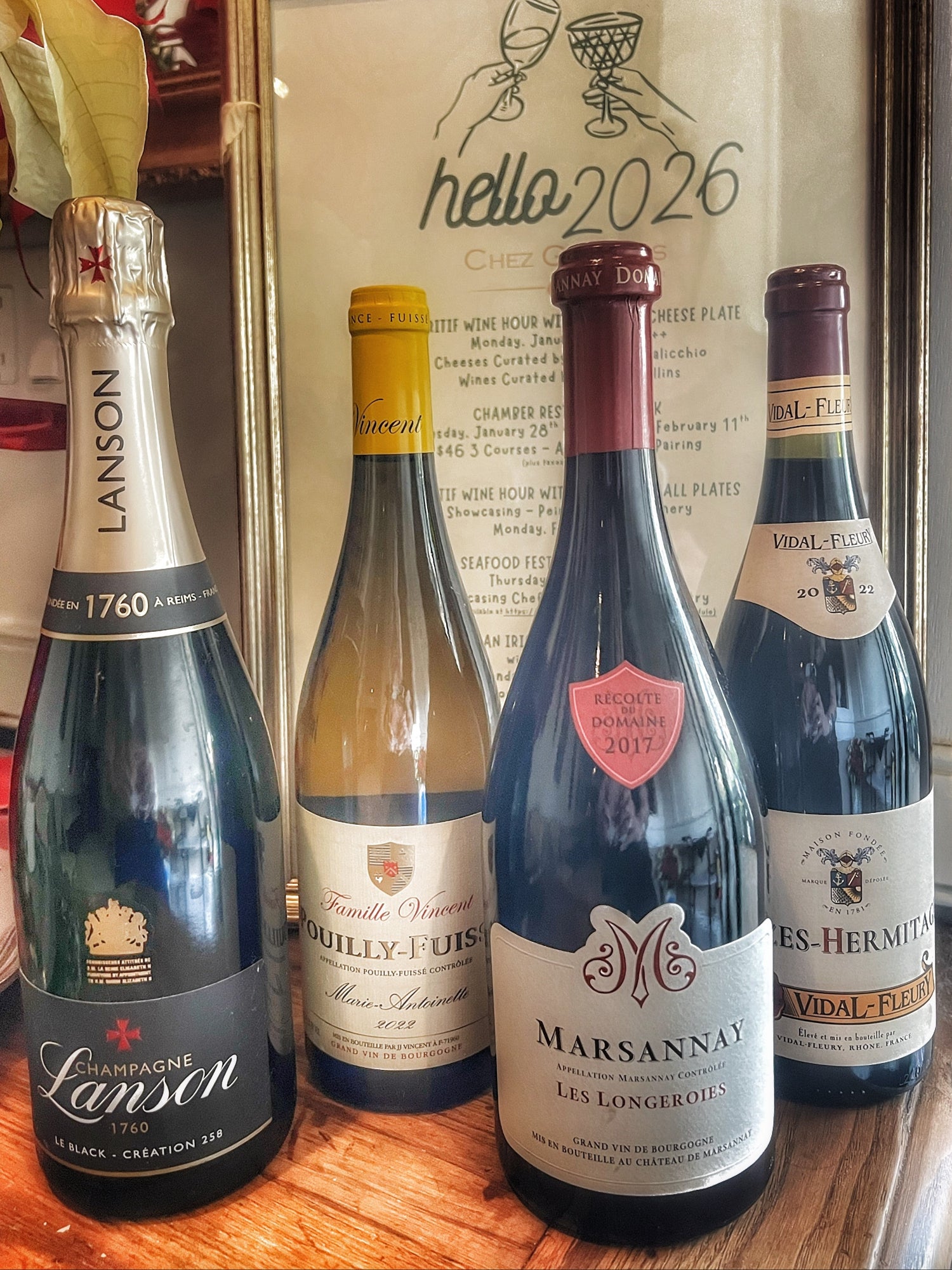 Four bottles of wine on a wooden surface with a decorative mirror in the background displaying 'hello 2026'.